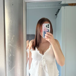 Vit linnetopp med volang och knyt - Supersöt vit linnetopp med volangkant vid urringningen och knyt framtill. Toppen är ärmlös och har en lätt rynkad struktur som ger en luftig känsla. Perfekt för varma dagar och enkel att styla med jeans eller kjol.