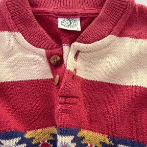 Randig tröja från Boomerang - Klassisk  stickad tröja från Boomerang i röd och vit med breda ränder och ett färgglatt mönsterband över bröstet. Tröjan har rund hals, knappar vid halsen och långa ärmar. Perfekt för dig som gillar retrovibbar och unika detaljer.