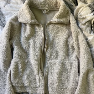 Beige teddyjacka med dragkedja - Mysig beige teddyjacka med hög krage och hel dragkedja framtill. Jackan har två stora fickor och är tillverkad i fluffigt fleece-material som ger en chill och avslappnad vibe. Perfekt för kalla dagar när du vill vara både varm och snygg. Från hm