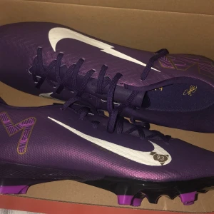 Lila Nike fotbollsskor med snörning - Säljer ett par lila Nike fotbollsskor med vita swoosh-detaljer och coola mönster i guld och lila. Skorna har snörning och platt sula med dobbar för bra grepp på planen. Perfekta för dig som vill sticka ut på fotbollsplanen!