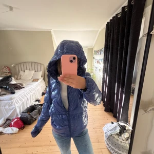 Blå glansig pufferjacka med huva - Säljer en blå, glansig pufferjacka med huva och dragkedja framtill. Jackan har en quiltad look och är perfekt för kalla dagar