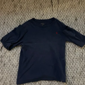 Mörkblå t-shirt från Polo Ralph Lauren - Klassisk mörkblå långarmad från Polo Ralph Lauren med röd broderad logga på bröstet. T-shirten har rund halsringning och långa ärmar. Tillverkad i mjuk bomull för en skön känsla och enkel stil. Har en litet hål under armen men det syns knappt.  