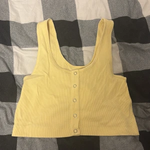 Gul ribbad linnetopp från Bik Bok - Snygg ljusgul croppad linnetopp från Bik Bok med ribbad struktur och tryckknappar framtill. Toppen är ärmlös och har en rundad halsringning. Perfekt för varma dagar och enkel att matcha med jeans eller kjol.