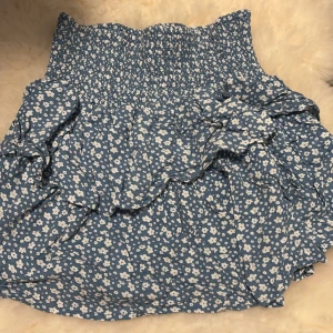 Blommig blå volangkjol från Hilt - Supersöt blå kortkjol från Hilt med vita små blommor och volangdetaljer. Kjolen har smock i midjan för en bekväm passform och är perfekt för vår och sommar. Lätt och luftig känsla, passar till sneakers eller sandaler.