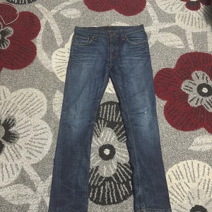 Blå jeans från Nudie Jeans, 31/32 - Säljer ett par klassiska blå jeans från Nudie Jeans med snygga slitningar och kontrastsömmar. Modellen har raka ben och ikoniska bakfickor med broderad våg. Jeansen är i bomull och har normal passform, perfekta för en avslappnad stil. Använd jätte lite