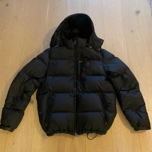 Svart dunjacka från Polo Ralph Lauren - Säljer en svart dunjacka från Polo Ralph Lauren med huva och dragkedja. Jackan har en klassisk pufferstil, två sidofickor med dragkedja och en liten röd logga på bröstet. Perfekt för kalla dagar och riktigt snygg till streetwear.