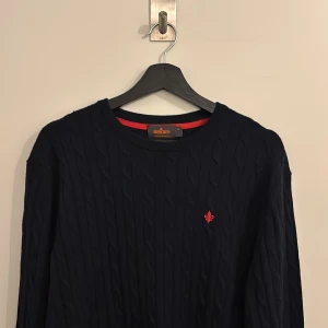Mörkblå kabelstickad tröja Morris - Kabelstickad mörkblå tröja från Morris i 100% merinoull. Klassisk rund halsringning och diskret röd logga broderad på bröstet. Perfekt för dig som gillar stilrena och tidlösa plagg med en modern touch. Storlek XL i tröjan men passar mer L enligt mig.