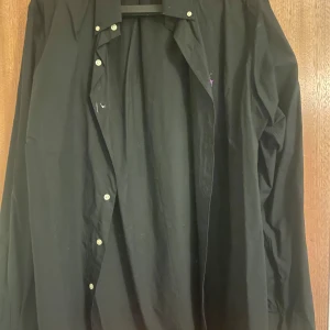 Svart skjorta Polo Ralph Lauren XL - Svart skjorta från Polo Ralph Lauren i slim fit-modell. Klassisk krage, vita knappar och lång ärm. Skjortan är tillverkad i bomull och har en stilren look som passar perfekt till jeans eller chinos.