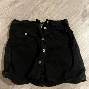 Svart kortkjol med knappar - Svart kortkjol från Replay.  Med stora framfickor och vita knappar längs hela framsidan. 