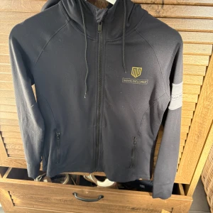 Tröja  från Maya Delorez  - Svart hoodie från Maya Delorez med gråa detaljer och logga på bröstet samt stor logga på ryggen. Tröjan har dragkedja framtill, huva med snörning och två fickor med dragkedja. Storlek S, original pris 1000kr.