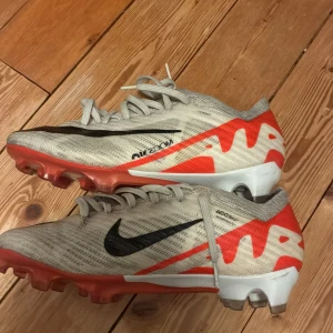 Nike Air Zoom Mercurial ELITE fotbollskor - Säljer ett par Nike Air Zoom Mercurial fotbollsskor i vit med röda och svarta detaljer. Skorna har snörning, svart swoosh-logga och en platt sula med dobbar för bra grepp på planen. Ovandelen är i syntetmaterial och designen är riktigt sportig och modern. Elite. Storlek 36,5. Använda 4 gånger.