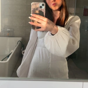 Vit klänning med puffärmar - Superfin vit klänning med puffiga långärmar och knappar framtill. Klänningen har en lätt strukturerad yta och markerad midja som ger en snygg siluett. Perfekt för studenten 