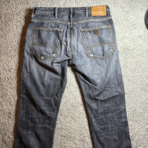 Emporio Armani grå jeans med patch - Snygga grå jeans från Emporio Armani med klassisk femficksdesign, kontrastsömmar och logga på bakfickan. Framtill finns en orange patch och slitning vid knät för en cool, avslappnad look. Tillverkade i jeansmaterial med rak passform. Den är lite sliten passar om du är 165-173. Inget är sönder men dem är bara användna