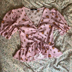 Rosa blommig blus  - Supersöt rosa blus från Bik Bok med orange blommönster, v-ringning och knyt i midjan. Blusen har volang nertill, puffiga korta ärmar och smockdetaljer vid ärmslut. Perfekt för dig som gillar romantisk och trendig stil🌸✨