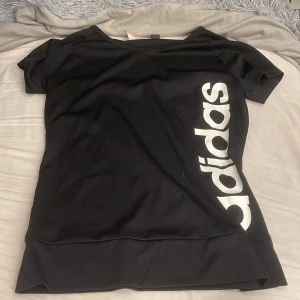 Svart Adidas t-shirt med logga - Svart t-shirt från Adidas med stor vit logga längs sidan. Klassisk rund halsringning och korta ärmar. Skön passform och tillverkad i mjukt material, perfekt för en sportig och avslappnad stil.