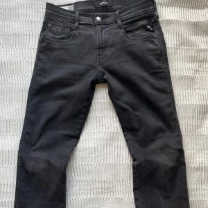 Svarta Replay Anbass jeans slim fit - Snygga svarta Replay Anbass jeans i slim fit-modell. Klassisk femficksdesign, svarta sömmar och diskret Replay-logga på fickan. Jeansen är tillverkade i stretchigt jeansmaterial för en bekväm passform och har en modern, smal siluett. Storlek 30/30. 