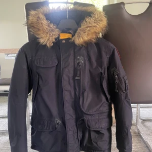 Svart Parajumpers jacka med fuskpäls - Säljer en svart Parajumpers jacka med huva och fuskpäls. Jackan har flera fickor, dragkedja och knappar framtill samt patch på ärmen. Perfekt för kalla dagar med sin varma och rejäla känsla. Klassisk streetstil med snygga detaljer och markerad gul rem vid halsen.