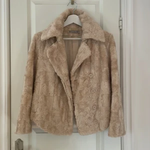 INTRESSEKOLL Beige pälsjacka från Kookai - INTRESSEKOLL på mysig beige pälsjacka från Kookai med stora slag och knäppning framtill. Jackan har lång ärm och en klassisk, lite boxig passform. Perfekt för dig som vill ha en cozy och trendig look under kyliga dagar.