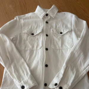 Vit overshirt från Jack & Jones S - Snygg benvit overshirt från Jack & Jones Premium med svarta knappar och två bröstfickor med lock. Skjortan är långärmad och gjord i mjuk bomull, perfekt att ha som lager över t-shirt. Klassisk krage och stilren design.