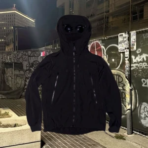 Svart Goggle Jacket från C.P. Company - Svart Goggle Jacket från C.P. Company med ikoniska goggles i huvan. Jackan har dragkedjor framtill och på fickorna, justerbar huva och ribbade muddar. Tillverkad i slitstark polyester, perfekt för dig som gillar streetwear och vill sticka ut.