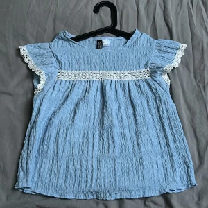 Blå blus med spets från H&M Divided - Supersöt ljusblå blus från H&M Divided med volangärmar och vit spetsdetalj över bröstet och vid ärmslut. Blusen har en krinklad struktur. Passar perfekt till de flesta tillfällen och är använd max 1 gång.