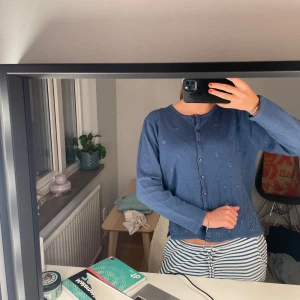 Blå broderad kofta från D&D Collection - Säljer en blå kofta från D&D Collection i storlek S. Koftan har broderade blommor och mönster framtill, lila knappar och rund halsringning. Den är stickad och har långa ärmar, perfekt för lager-på-lager under kyliga dagar.