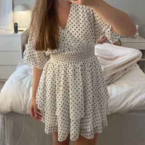 Vit prickig kortklänning med volang - Superfin vit kortklänning med svarta prickar och volangdetaljer. Klänningen har v-ringad hals, smockad midja och luftiga korta ärmar. Perfekt för sommarens alla tillfällen, riktigt söt och trendig look.