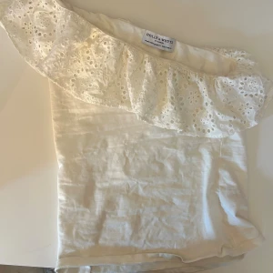 Vit offshoulder topp Holly & Whyte - Supersöt vit offshoulder topp från Holly & Whyte med bred volang i broderad spets. Toppen är ärmlös och har en romantisk känsla med sin ljusa färg och fina detaljer. Perfekt för varma dagar och somriga outfits.