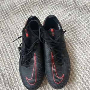 Nike Phantom GT2 fotbollsskor svart/röd - Säljer ett par Nike Phantom GT2 fotbollsskor i svart med röda detaljer och Nike-logga. Skorna har snörning, rund tå och platt sula med dobbar för bra grepp på planen. Ovandelen är i syntetmaterial med textur för bättre bollkontroll. Perfekta för dig som vill sticka ut på fotbollsplanen.