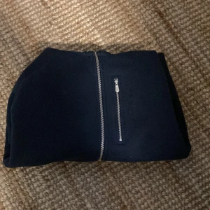 Svart hoodie från Sail Racing - Säljer en svart hoodie från Sail Racing med hel dragkedja och en extra dragkedjeficka på bröstet. Tröjan har huva med snörning och är tillverkad i mjukt material, perfekt för kyliga dagar. Enkel och stilren design med diskret logga på bröstet.