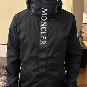 Marinblå vindjacka från Moncler - McKinley - Snygg blå vindjacka från Moncler med stor logga framtill och patch på ärmen. Jackan har huva med dragsko, vita dragkedjor och är tillverkad i glansig polyester. Perfekt för dig som vill ha en stilren och exklusiv look. Obetydligt använd, märkt med storlek 2, passar S. Nfc fungerar inte. 
