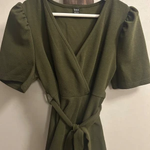 Olivgrön omlottklänning från SHEIN - Olivgrön kortklänning från SHEIN med omlottmodell och knytband i midjan. Klänningen har korta puffärmar och v-ringad hals. Materialet känns mjukt och lätt, perfekt för en chill look.