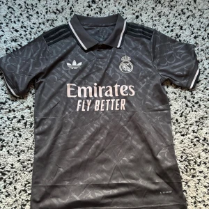 Real Madrid Adidas fotbollströja S - Snygg mörkgrå Real Madrid fotbollströja från Adidas med klassisk krage och vita detaljer. Tröjan har korta ärmar, diskret mönster i tyget och broderade loggor. Materialet är lätt och andas, perfekt för träning eller match.