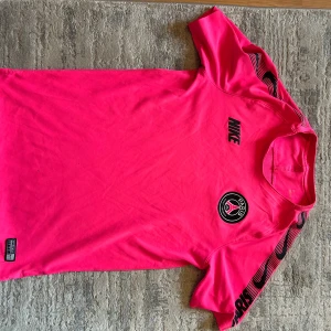 Paris Saint-Germain Nike fotbollströja rosa - Säljer en riktigt snygg Paris Saint-Germain fotbollströja från Nike i stark rosa färg. Tröjan har korta ärmar med svarta Nike-loggor längs axlarna och PSG-emblem på bröstet. Tillverkad i lätt och ventilerande material, perfekt för träning eller match. Tröjan har 2-3 små hål därav priset 