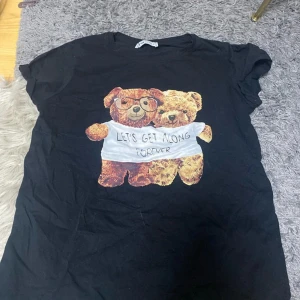 Svart t-shirt med nalletryck från B Sister - Svart t-shirt från B Sister med korta ärmar och ett gulligt tryck av två nallar på framsidan. Nallarna har en gemensam tröja med texten 'LET'S GET ALONG FOREVER'. Skön och mjuk bomull, perfekt för en chill look.