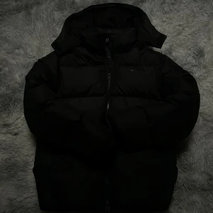 Svart pufferjacka Tommy Hilfiger - Snygg svart pufferjacka från Tommy Hilfiger med huva och dragkedja. Jackan har diskret logga på bröstet och ett patch på ärmen. Perfekt för kalla dagar. Den är köpt ifrån kids brand store och har nu pris 1800. Och är storlek 164cm
