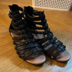 Svarta gladiatorsandaler med nitar - Snygga svarta gladiatorsandaler med flätade och släta remmar, små silverfärgade nitar och dragkedja bak. Öppen tå och platt sula gör dem perfekta för sommaren. Tillverkade i syntetmaterial och har en cool, edgy vibe.