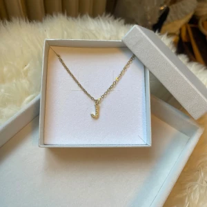 Guldhalsband med glittrig J-berlock - Snyggt halsband i guld med en glittrande berlock formad som bokstaven J. Kedjan är tunn och elegant, och berlocken är täckt av små gnistrande stenar som ger ett lyxigt intryck. Perfekt accessoar för dig som vill bära din initial eller ge bort som personlig present.