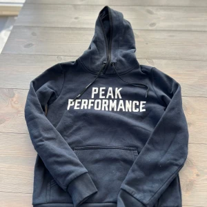 Mörkblå hoodie från Peak Performance - Säljer en mörkblå hoodie från Peak Performance i storlek XS. Tröjan har stor huva med snörning, stor vit logga framtill och en klassisk känguruficka. Mjuk insida och ribbade muddar vid ärmslut och nederkant. Perfekt för chill dagar eller när du vill vara extra bekväm.