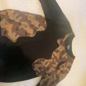 Fendi brun och svart sweatshirt - Cool svart sweatshirt från Fendi med bruna och beige camouflageinspirerade partier och FF-logga över hela mönstret. Tröjan har rund halsringning och långa ärmar. Perfekt för dig som vill sticka ut med designerstil.