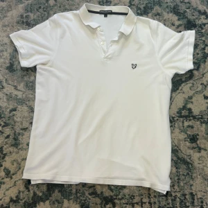 Vit piké från Lyle & Scott - Vit pikétröja från Lyle & Scott i storlek L men passar som M. Klassisk design med krage, knappar och broderad logga på bröstet. Tillverkad i mjuk bomull och har korta ärmar. Perfekt för en clean och stilren look.