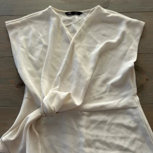 Vit omlottopp med knyt från Zara - Säljer en stilren vit omlottopp från Zara i storlek M. Toppen har ärmlös design, v-ringning och snygg knytning i midjan. Tillverkad i ett lätt och mjukt polyestertyg som ger ett elegant fall. Perfekt för dig som gillar minimalistisk och clean stil.