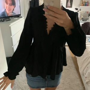 Svart blus med spets och v-ringning - Säljer en svart blus med lång ärm och v-ringning med fina spetsdetaljer längs kanten. Blusen har en lös passform och lätt volang vid ärmslut och nederkant. Perfekt att styla med jeanskjol eller byxor.