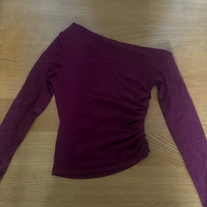 Vinröd one shoulder topp - Säljer en vinröd långärmad topp med one shoulder-design. Toppen har en tight passform och är tillverkad i ett mjukt, stretchigt material som sitter snyggt på kroppen. Perfekt för dig som vill ha en stilren men ändå unik look.