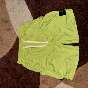 Limegröna Stone Island shorts - Säljer ett par limegröna shorts från Stone Island med elastisk midja och vit snörning. Shortsen har en klassisk patch på benet och praktiska fickor med dragsko. Perfekta för sommaren och riktigt snygga till streetwear-stilen.