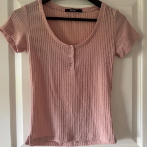 Rosa ribbad topp från Bik Bok - Säljer en ljusrosa ribbad topp från Bik Bok i storlek S. Toppen har korta ärmar, rund halsringning och knappar framtill. Materialet är mjukt och stretchigt, perfekt för en avslappnad look.