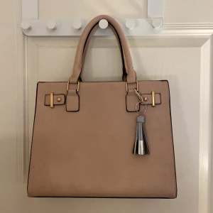 Beige handväska med tofsdetalj - Stilren beige handväska i skinnimitation med guldiga metalldetaljer och en snygg tofs i silver och grått. Väskan har två handtag, dragkedja baktill och flera fack inuti för smart förvaring. Perfekt för dig som gillar klassisk och chic look.