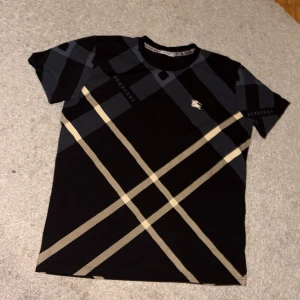 Svart rutig Burberry t-shirt  - Helt ny snygg svart t-shirt från Burberry med klassiskt rutigt mönster i beige och grått. T-shirten har korta ärmar, rund hals och broderad Burberry-ryttare på bröstet. Perfekt för dig som gillar stilrena designer och ikoniska loggor. Storlek är L men passar mer som M. Först till kvarn!!!