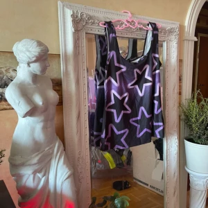 Top No°2 - Coolt svart linne med stora lila stjärnor över hela tyget. Modellen är ärmlös med breda axelband och rundad halsringning. Perfekt för dig som gillar statement-plagg och vill sticka ut lite extra. Materialet känns lätt och luftigt.