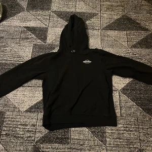 Svart hoodie från Biltema i storlek L barn - Svart hoodie från Biltema med liten vit logga på bröstet. Tröjan har huva med snörning och är tillverkad i en mjuk bomulls- och polyestermix. Klassisk passform med långa ärmar och ribbade muddar. Perfekt för chill dagar eller när du vill ha en enkel look.